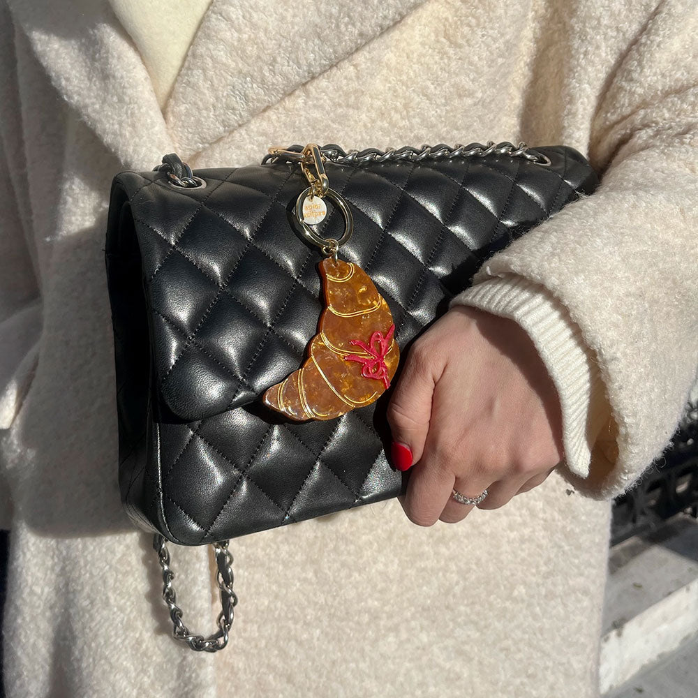 woman in a white coat holding a black quilted handbag with a brown croissant bag charm attached