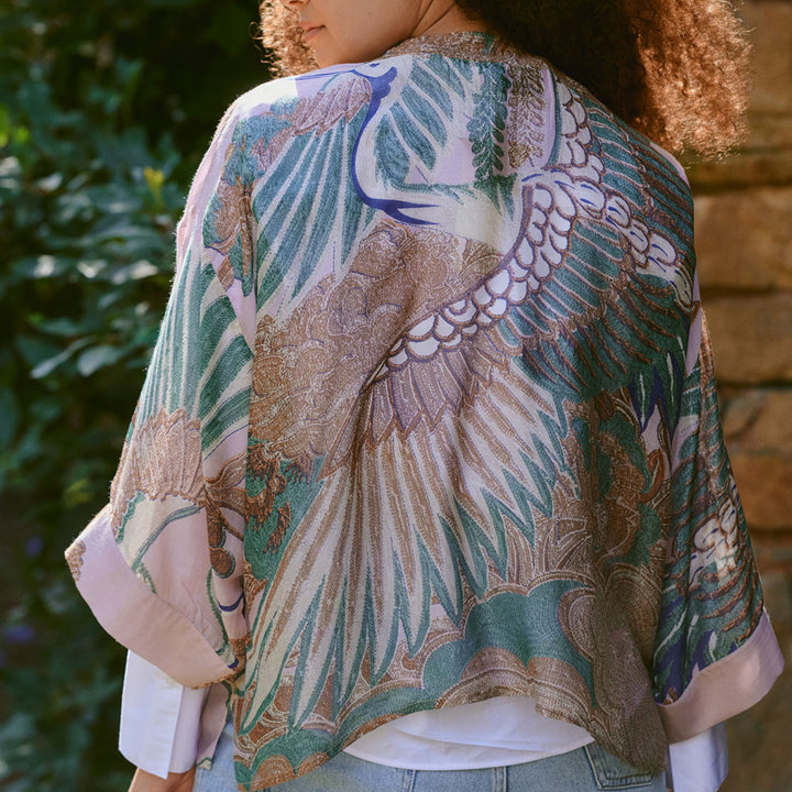 women's short kimono top featuring a design of cranes in green, brown & blue on a pale pink base