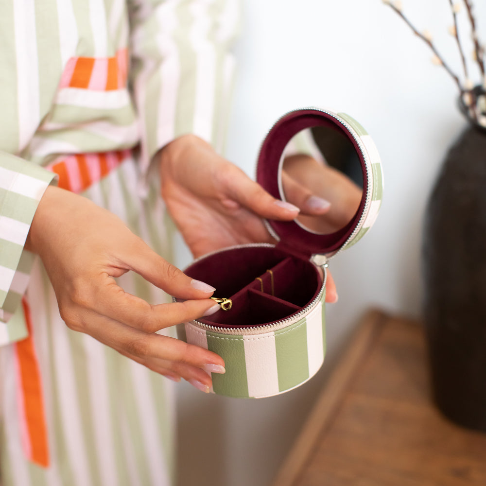 A person holds an open round jewelry box with a mirror on the inside lid. The box has a green and white striped exterior and a burgundy interior with a small gold ring holder.