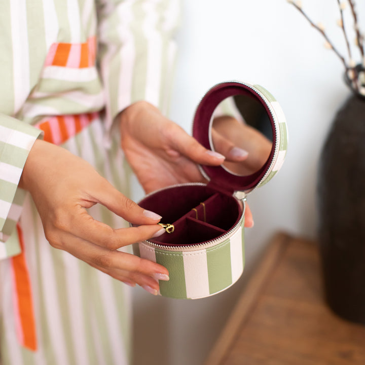 A person holds an open round jewelry box with a mirror on the inside lid. The box has a green and white striped exterior and a burgundy interior with a small gold ring holder.