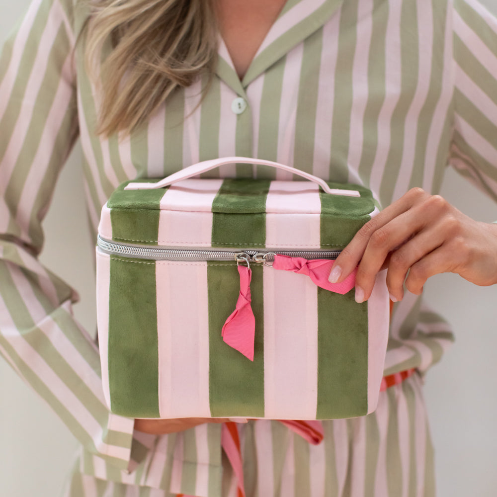 A woman in a green and white striped pajama shirt holds a matching green and white striped cosmetic bag with a pink zipper pull and pink ribbon accent.