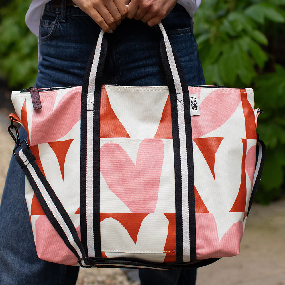 A person outdoors holding a large tote bag with a pink, orange, and white geometric heart pattern. The bag has black and white striped handles and a matching shoulder strap.