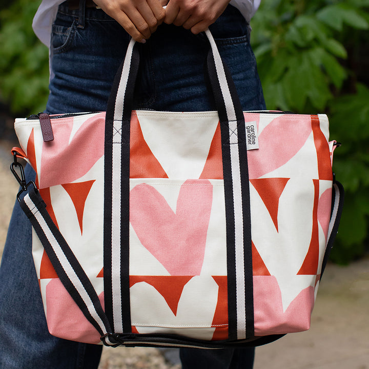 A person outdoors holding a large tote bag with a pink, orange, and white geometric heart pattern. The bag has black and white striped handles and a matching shoulder strap.