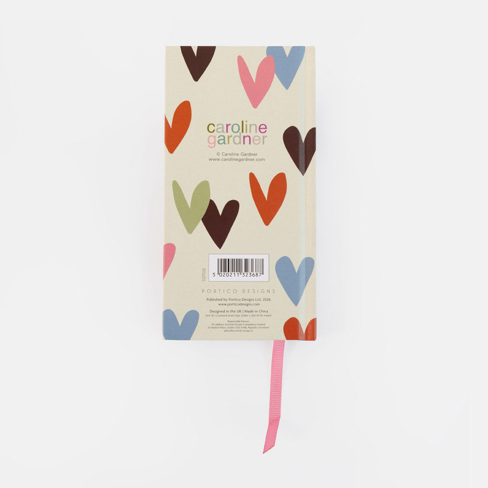 A Carolina Gardner pocket design notebook with a cream cover decorated with colorful heart illustrations, featuring a pink ribbon bookmark, shot against a plain white background.