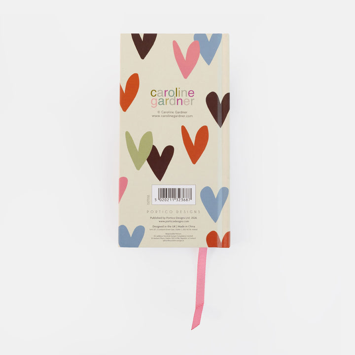 A Carolina Gardner pocket design notebook with a cream cover decorated with colorful heart illustrations, featuring a pink ribbon bookmark, shot against a plain white background.