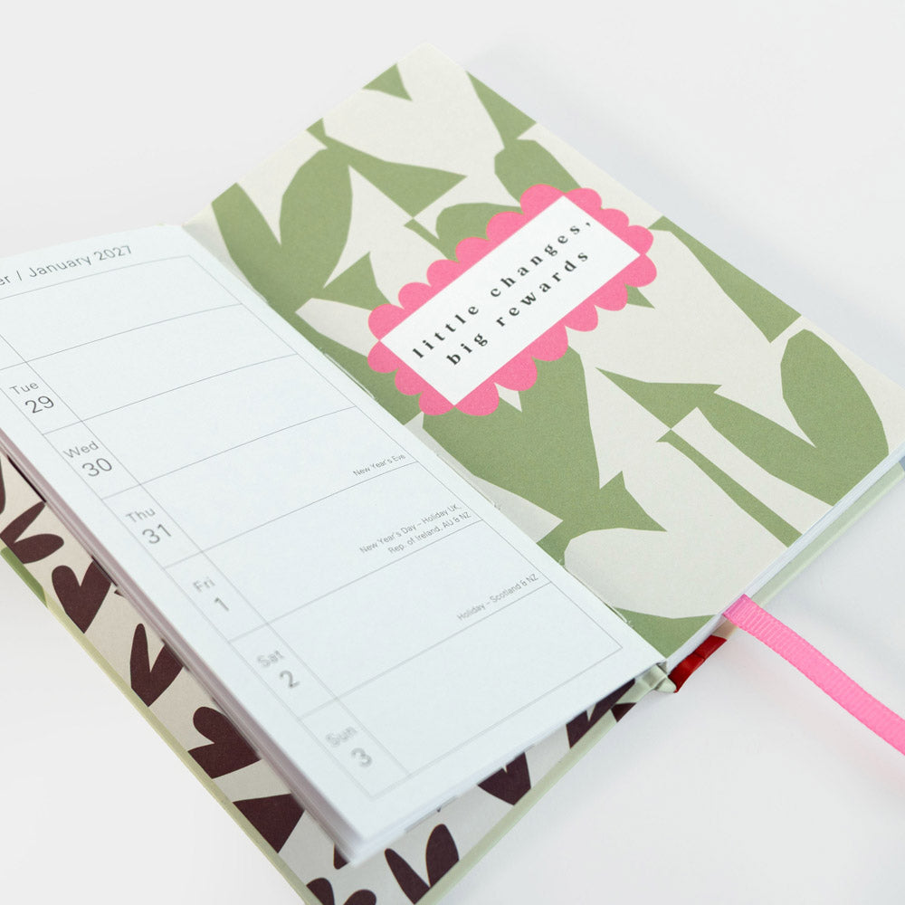 An open planner or journal with a green and white hearts pattern cover, a pink ribbon bookmark, and a white weekly page spread out on top. A pink label on the cover reads 'little changes, big rewards.