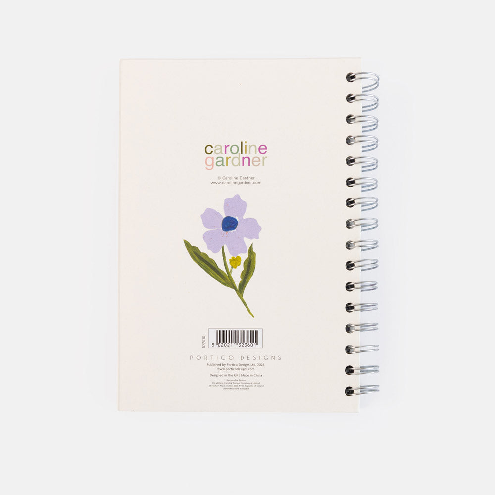 A spiral-bound notebook titled 'caroline gardener' on a clean white background. The cover is cream-colored with a purple flower illustration, green leaves, and a small yellow accent, featuring silver wire binding on the right side.