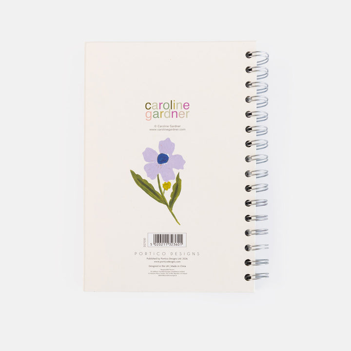 A spiral-bound notebook titled 'caroline gardener' on a clean white background. The cover is cream-colored with a purple flower illustration, green leaves, and a small yellow accent, featuring silver wire binding on the right side.