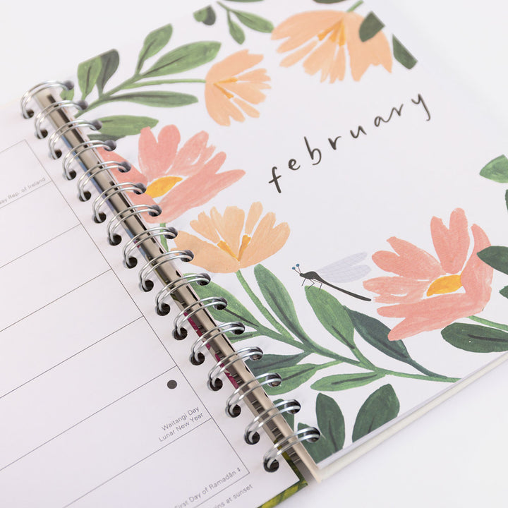 A spiral-bound planner or journal shown open on a white background, featuring a floral cover design with peach flowers, green leaves, and the handwritten word 'february'.