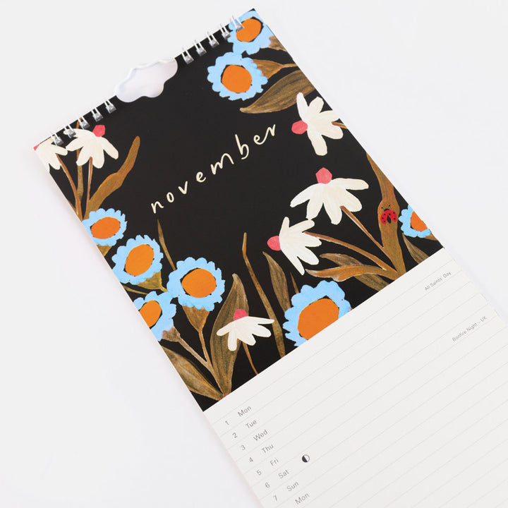 A spiral-bound November calendar with a black cover decorated with white, light blue, and orange flowers, plus a lined writing page visible at the bottom on a white background.