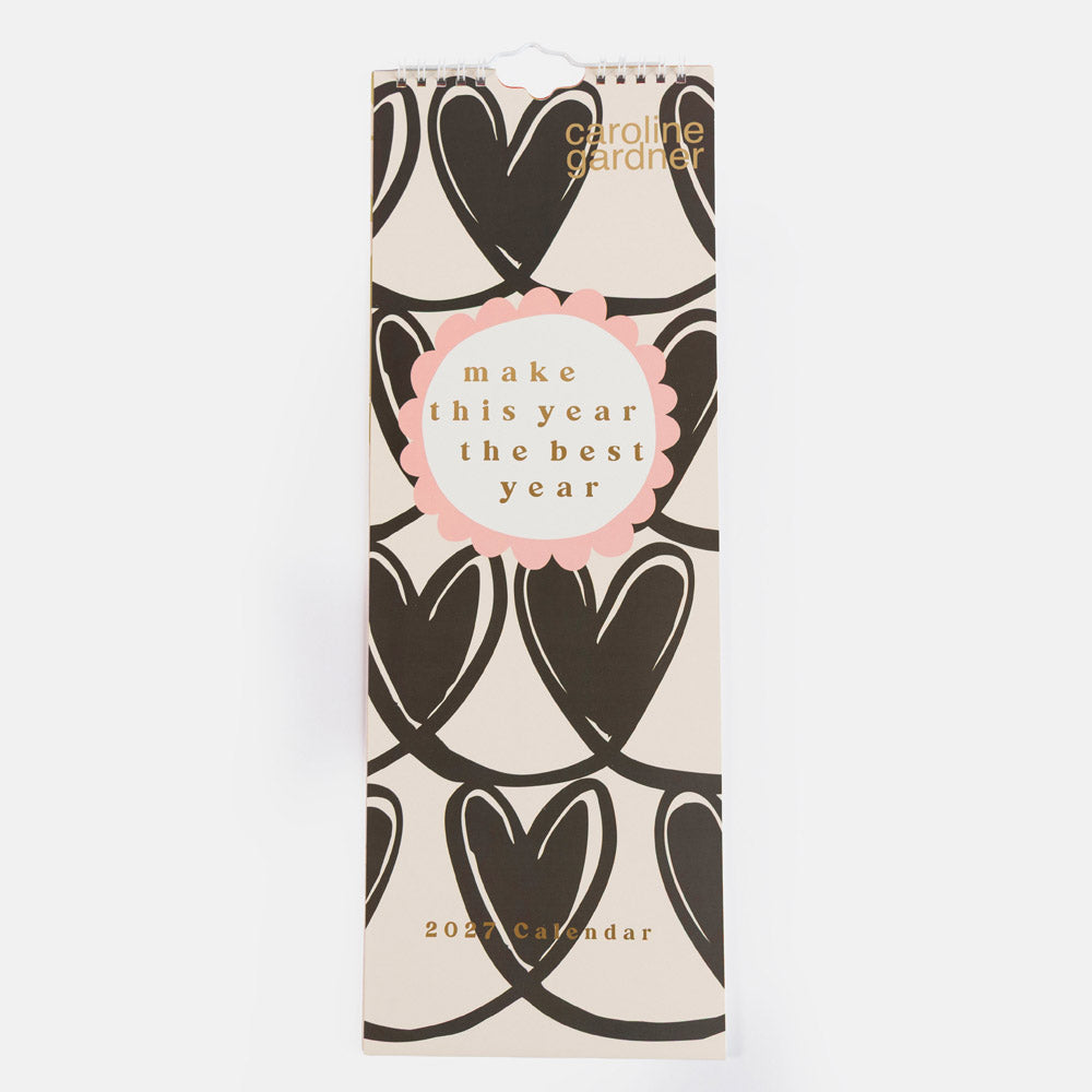 A tall, narrow 2027 spiral-bound calendar by Caroline Gardner, featuring a cream background with a repeating dark brown heart pattern and a central pink circular badge with the text 'make this year the best year'.