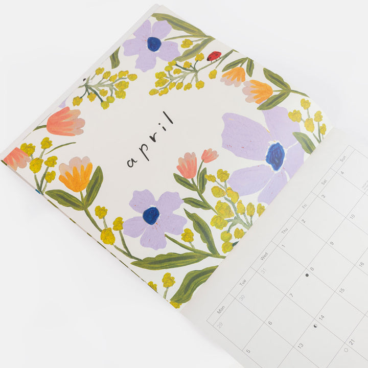 A floral monthly calendar page for April, featuring a white cover decorated with purple, orange, yellow, and green flowers, with a partial view of the printed calendar grid on the right.