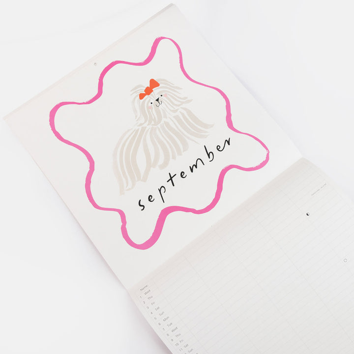 A close-up of a white calendar page featuring a decorative hand-drawn illustration of a long-haired dog inside a pink outline, with the word 'september' written in black script and an orange bow accent.
