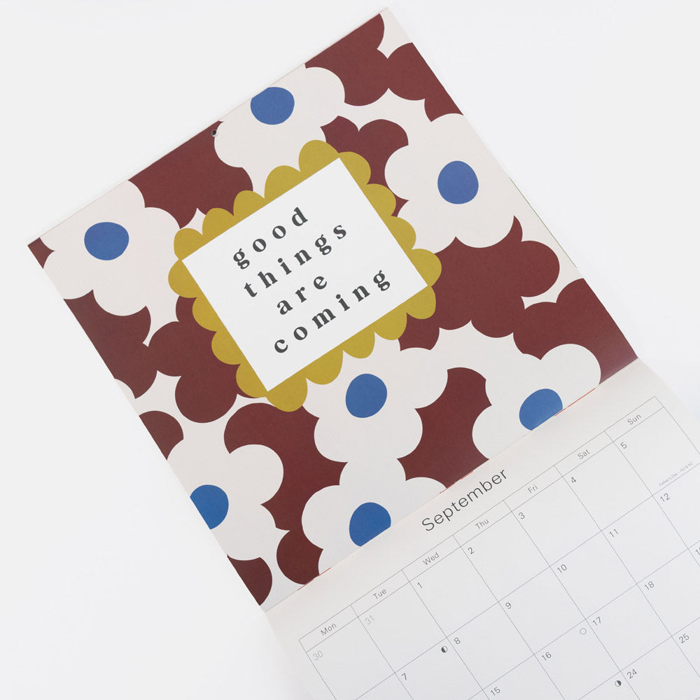 A square desk calendar with a retro floral pattern in brown, white, and blue, featuring a central white label with the text 'good things are coming' in a scalloped yellow border. The calendar is shown partially open, revealing a September calendar page on a clean white background.