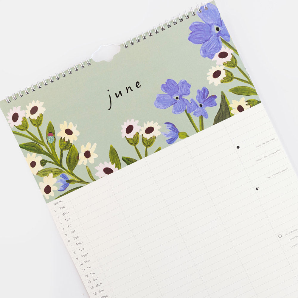 A spiral-bound monthly calendar page for June with a floral border design in lavender, white, and green on a pale green background, photographed on a clean white surface.