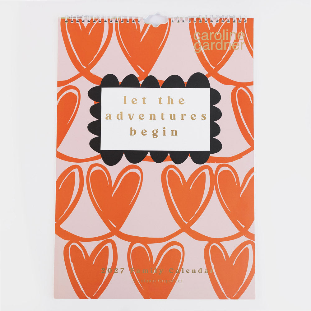 A spiral-bound Caroline Gardner 2027 yearly calendar featuring a pink background with orange heart patterns and a central white label with the text 'let the adventures begin'.