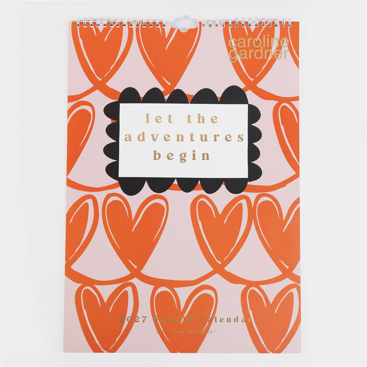 A spiral-bound Caroline Gardner 2027 yearly calendar featuring a pink background with orange heart patterns and a central white label with the text 'let the adventures begin'.