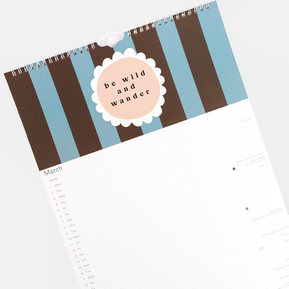 A spiral-bound calendar shown at an angle on a light gray background. The top cover has vertical stripes in dark brown, light blue, and white, with a peach circle in the center reading 'be wild and wander.