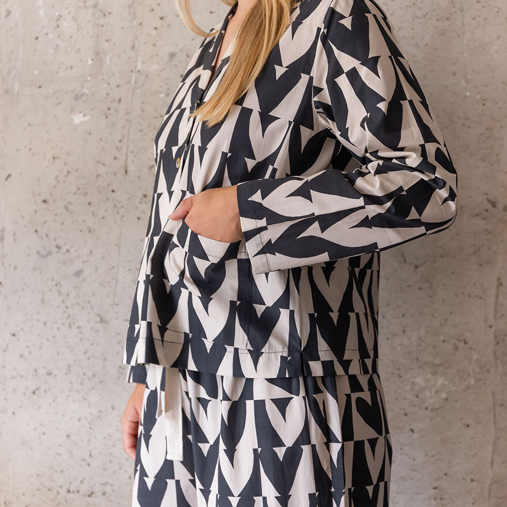 A woman wearing a matching long-sleeve button-front pyjama shirt and wide-leg pyjama pants set in a black and off-white hearts pattern, standing against a textured concrete wall.
