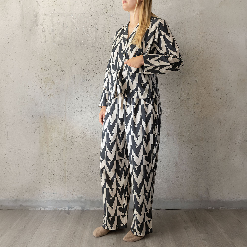 A woman wearing a matching long-sleeve button-front pyjama shirt and wide-leg pyjama pants set in a black and off-white hearts pattern, standing against a textured concrete wall.