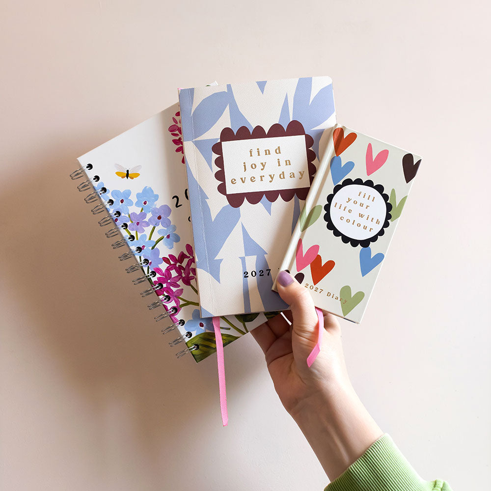 A hand holding three small decorative diaries fanned out against a plain light beige background. The covers feature pastel floral and heart patterns with handwritten-style quotes such as 'find joy in everyday' and 'live your life with joy.
