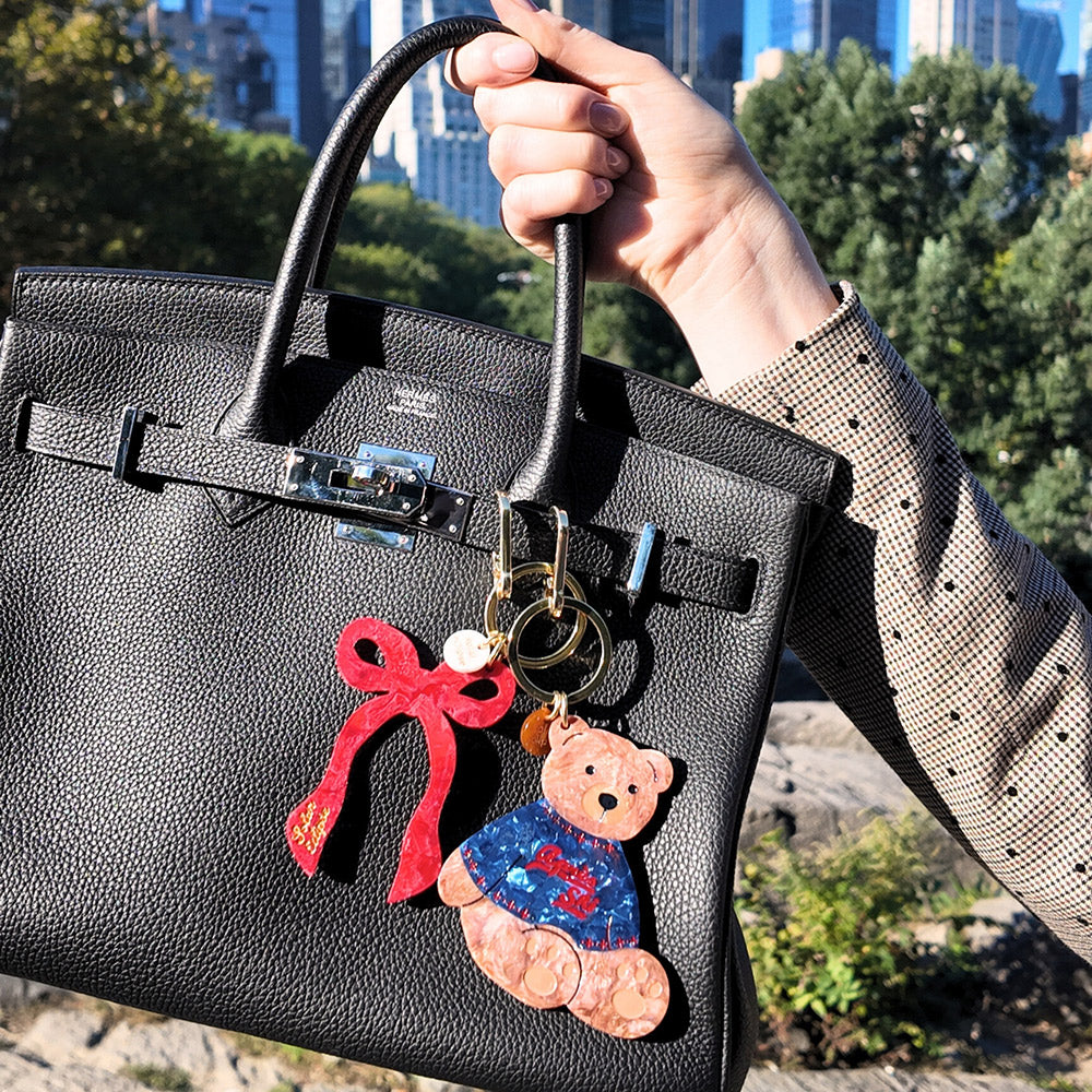 Black handbag with teddy bear and red bow keychain held by a person outdoors.