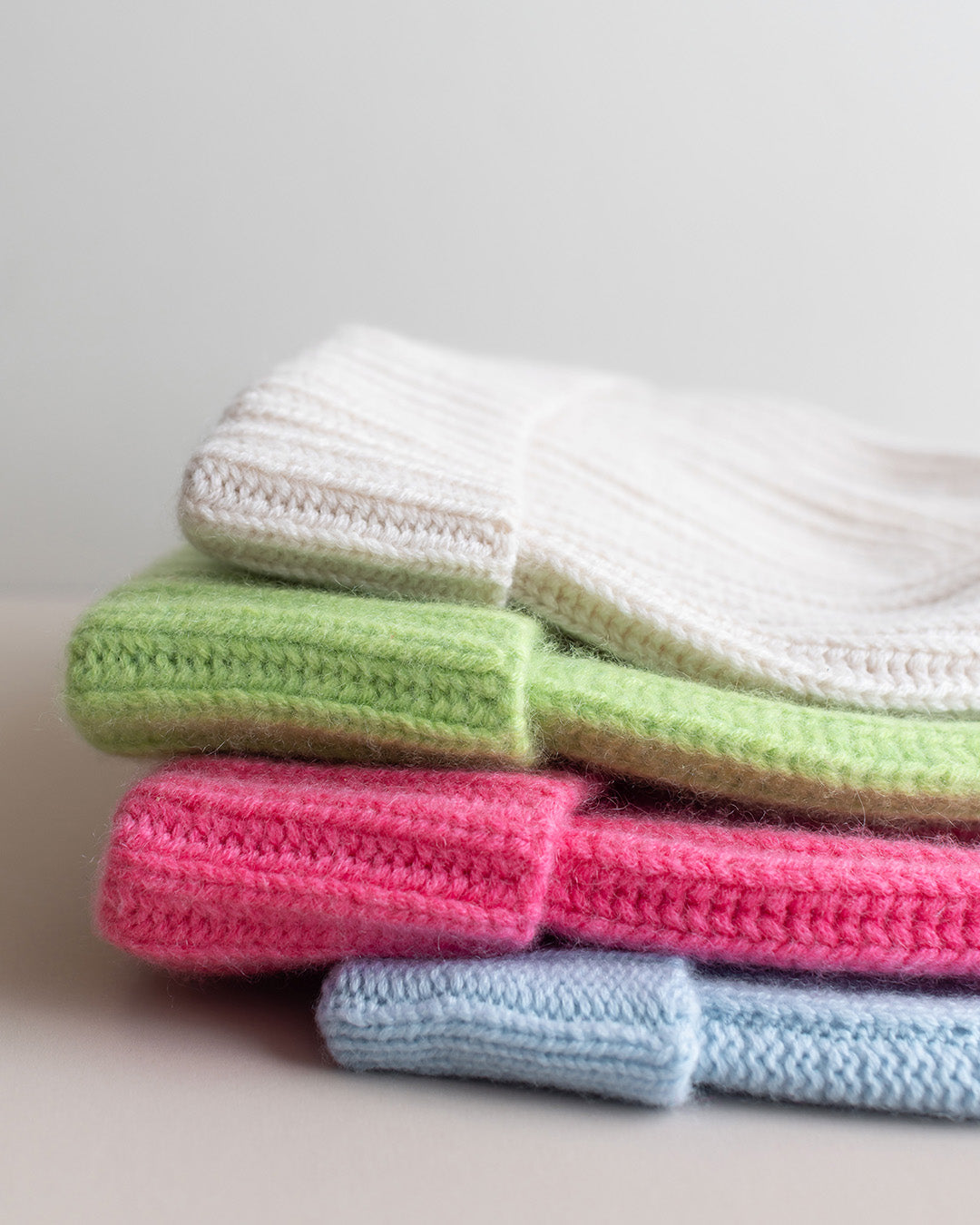 a stack of 4 colourful wool hats in cream, bright green, pink and light blue against a light grey background