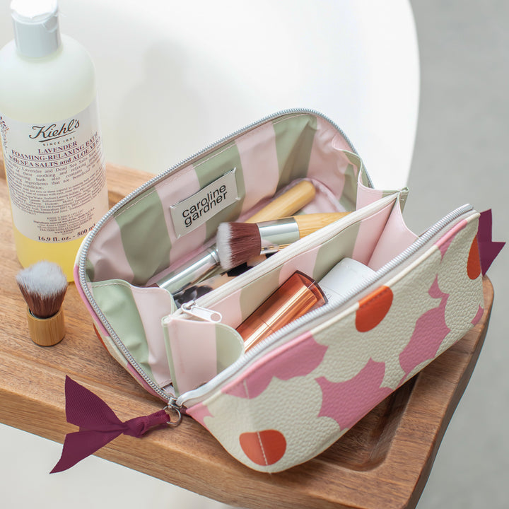 A cosmetic bag with a pink and orange patterned exterior and a pastel interior, shown open on a wooden tray with makeup brushes and beauty products inside. A Kiehl's bottle is visible in the background.