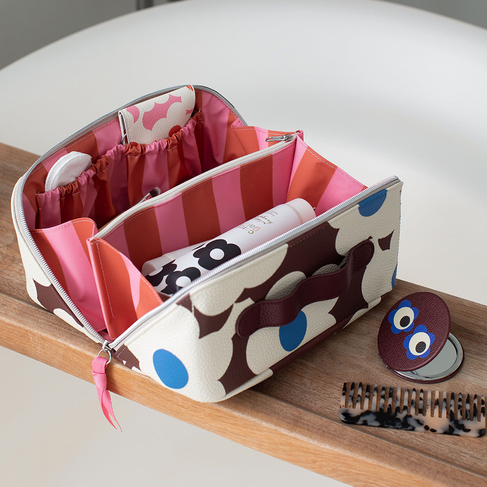 A zippered cosmetic organizer bag with a brown, pink, and blue flower-print pattern, shown open on a wooden surface with various beauty items inside.
