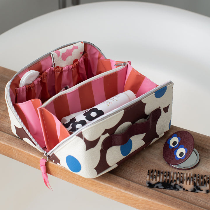 A zippered cosmetic organizer bag with a brown and cream flower print pattern, blue polka dots, and pink striped inner compartments, shown open on a wooden surface next to a small round mirror and a hairbrush.