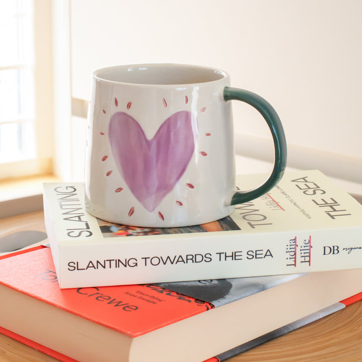 A white ceramic mug with a dark green handle and a purple heart design, resting on a stack of books in a bright indoor setting.