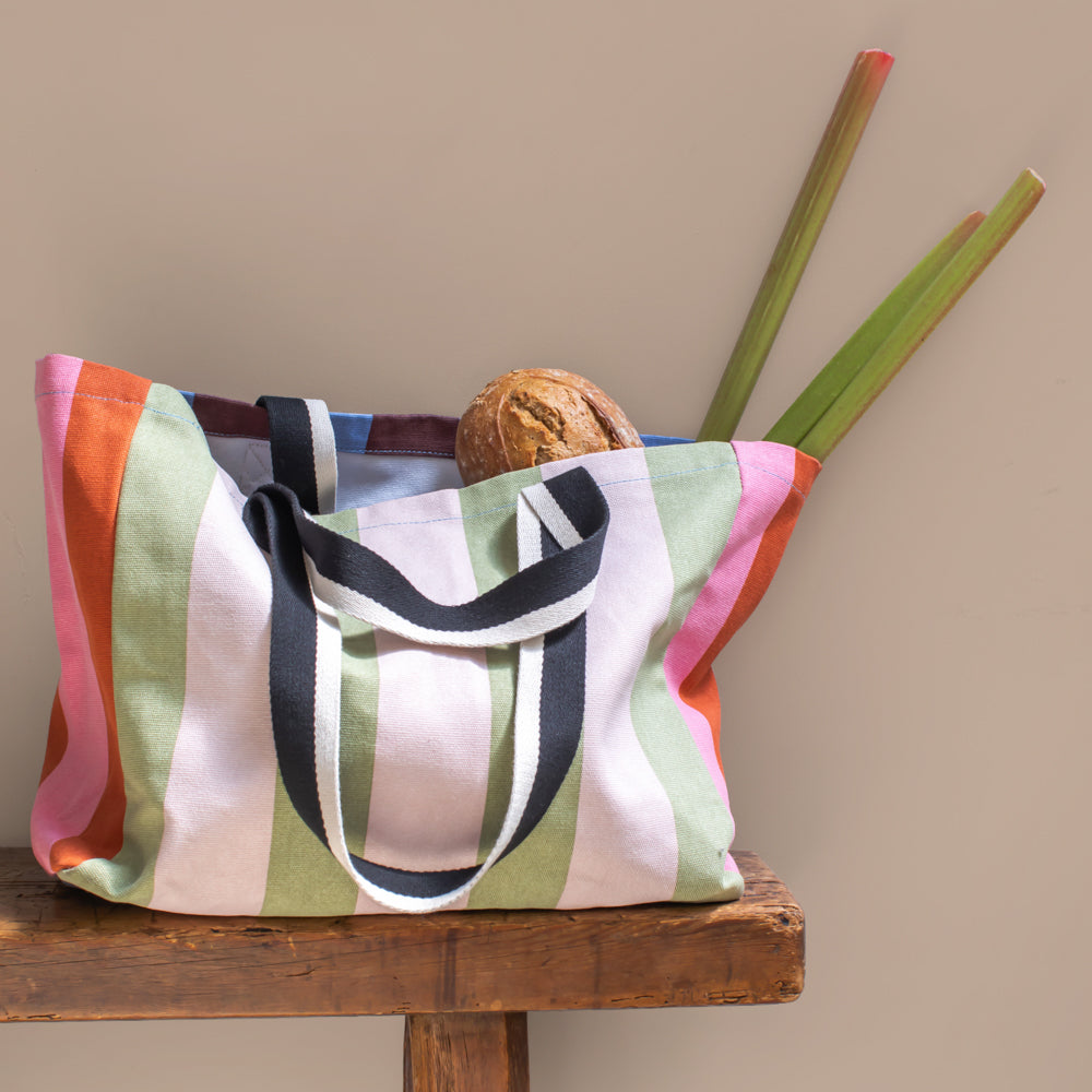 a colourful stripey tote bag with pink and green stripes on one side, and orange and pink green stripe on the other, sat on a wooden bench