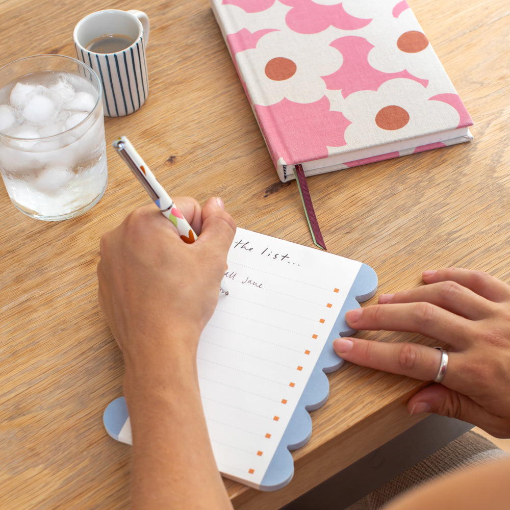a person writing on a blue scallop edged list pad on a wooden desk with a pink floral notebook, espresso cup and glass of water
