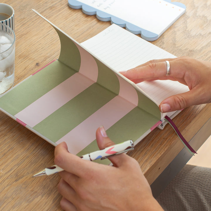a person holding a colourful heart design pen and opening a lined notebook with a pink and green striped inside cover on a wooden surface