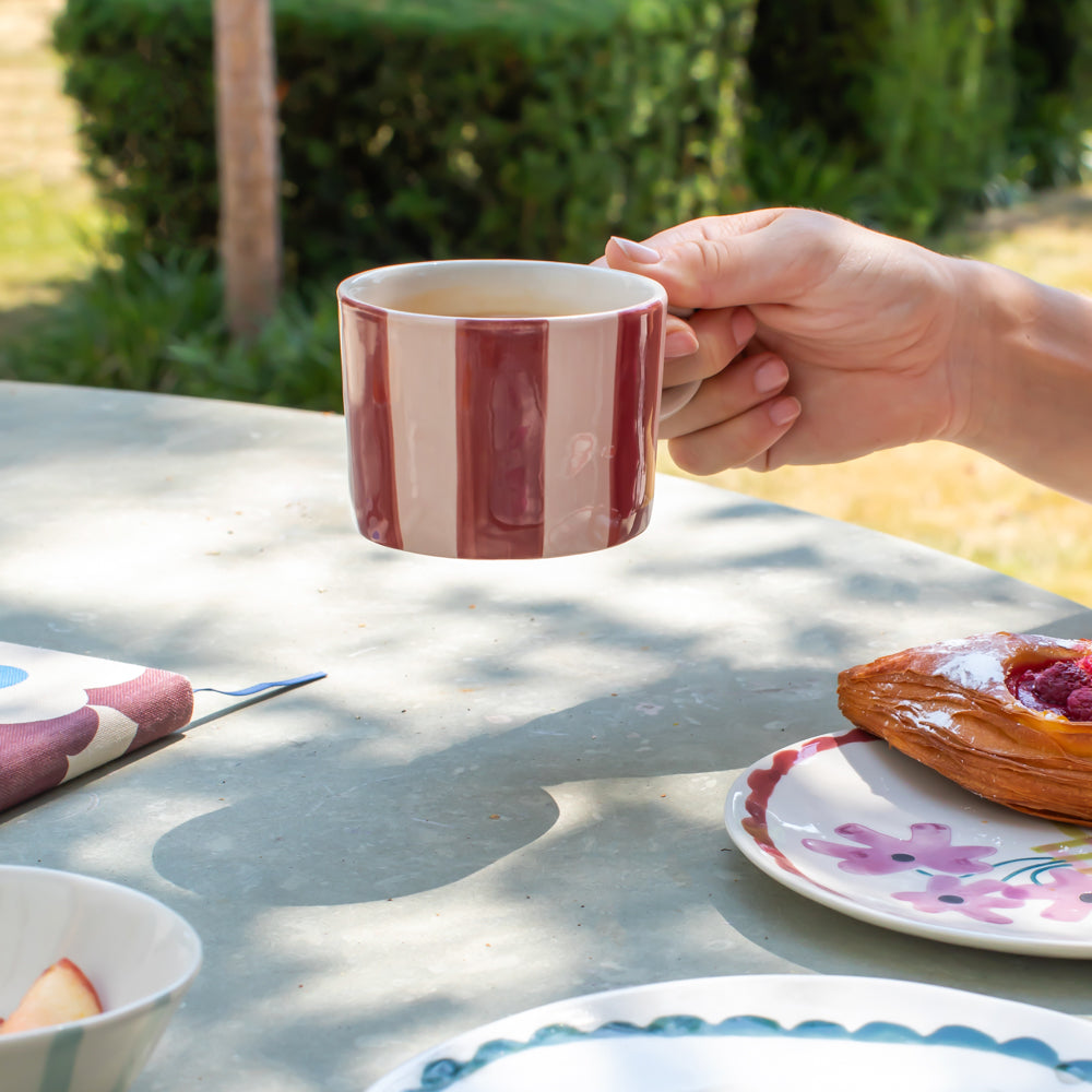 A hand holds a striped ceramic mug with vertical bands of deep red and cream, set on an outdoor patio table with plates, food, and greenery in the background.