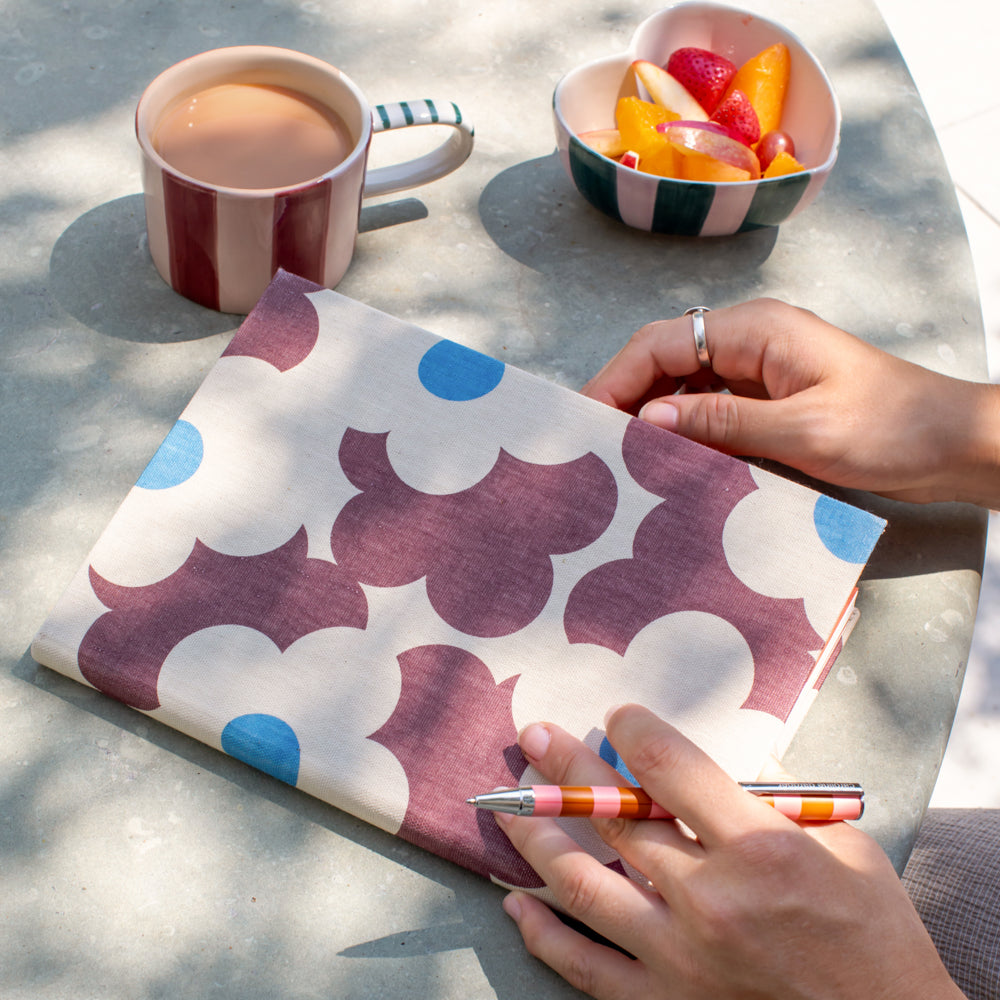 a person holding an orange and pink striped pen with a purple and blue floral design notebook, cup of tea and small bowl of chopped fruit