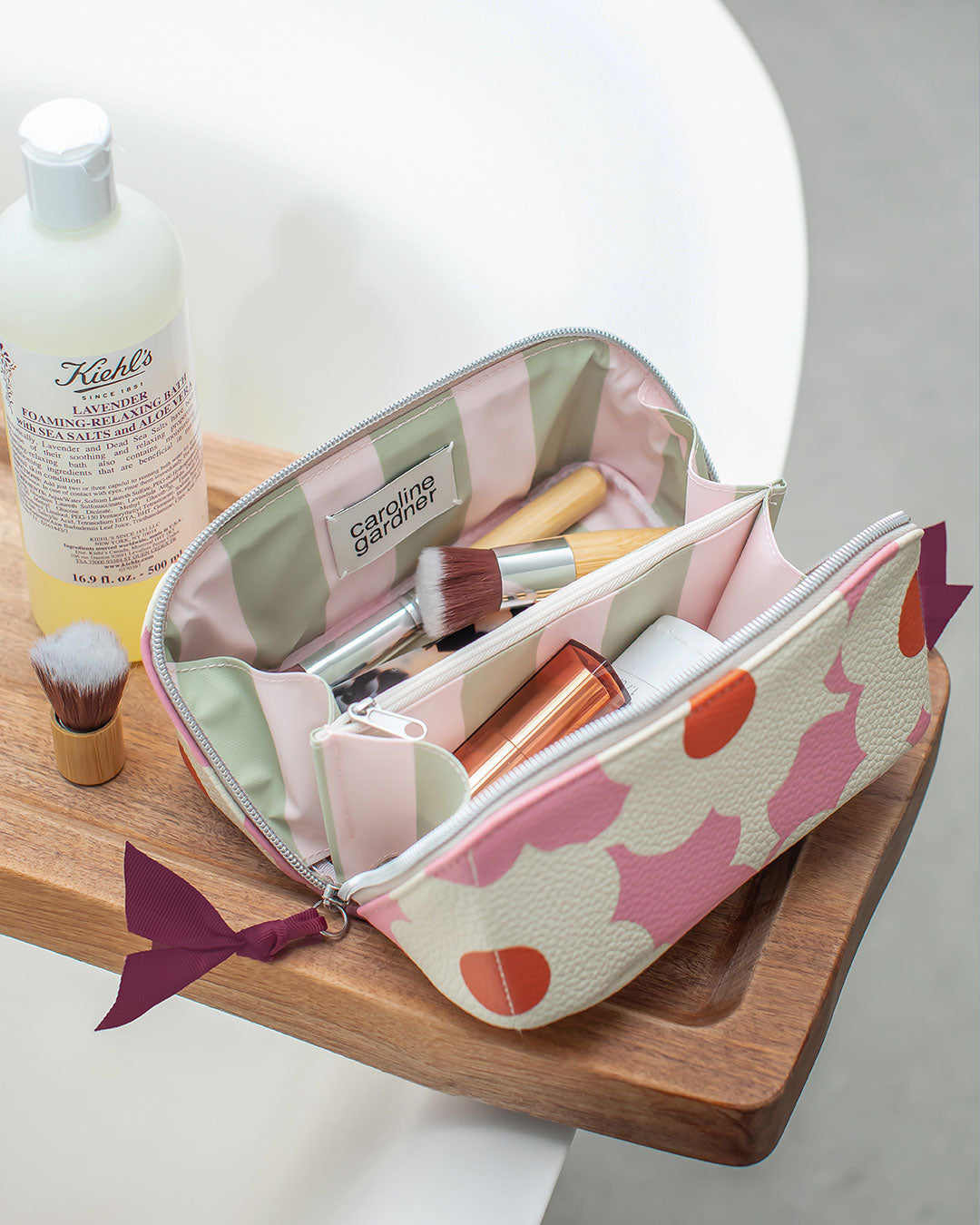 A pastel makeup bag with pink and orange abstract shapes sits open on a wooden tray, filled with cosmetics. A Kiehl's bottle is visible in the background, and the scene is styled with a white curved surface.