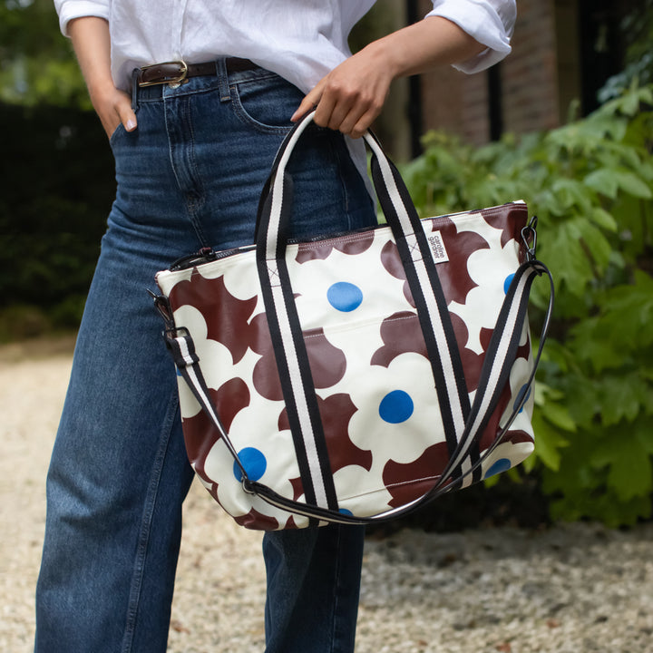 A person wearing blue jeans and a white shirt carries a large patterned tote bag outdoors. The bag has a cream base with brown abstract shapes and bright blue polka dots, plus black and white striped handles and shoulder strap.