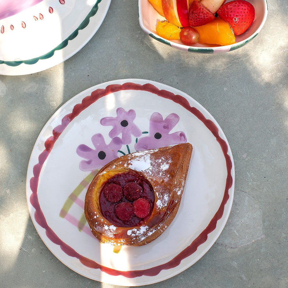 A close-up overhead shot of a plated pastry on a white ceramic plate with a scalloped red rim, floral print, and green stems. The plate sits on a gray surface with dappled sunlight, alongside a bowl of sliced fruit.