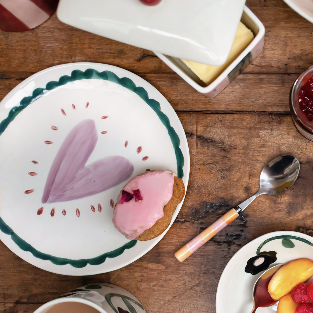 A styled breakfast scene on a rustic wooden table featuring a white ceramic plate with a green scalloped border and a purple heart design, holding a pink-frosted pastry. A pink-and-cream handled spoon, butter dish, jam, and other breakfast items are arranged around it.