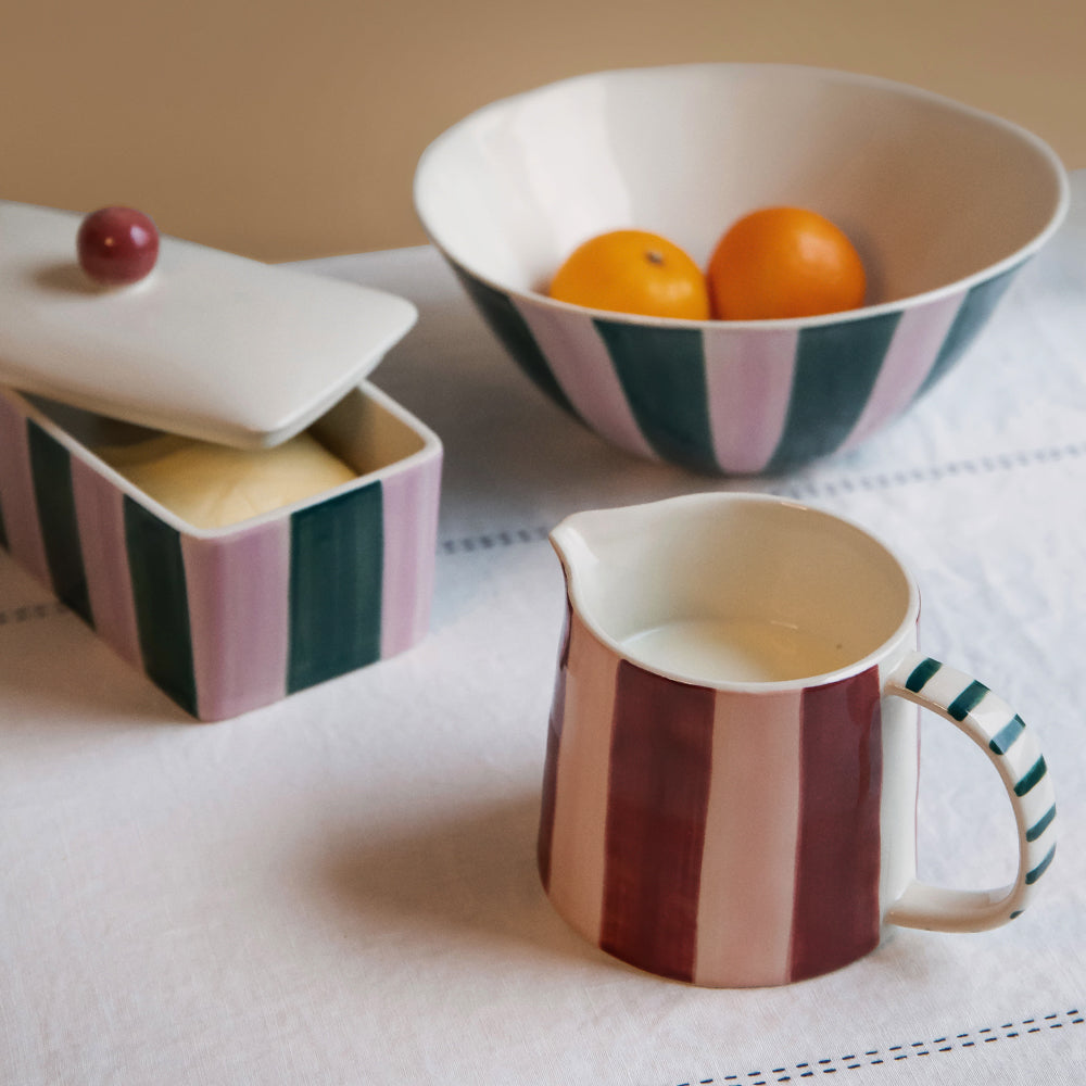 A coordinated set of ceramic tableware including a lidded butter dish, a bowl with oranges, and a creamer pitcher, all featuring vertical pink, dark green, and cream stripes on a white tablecloth.