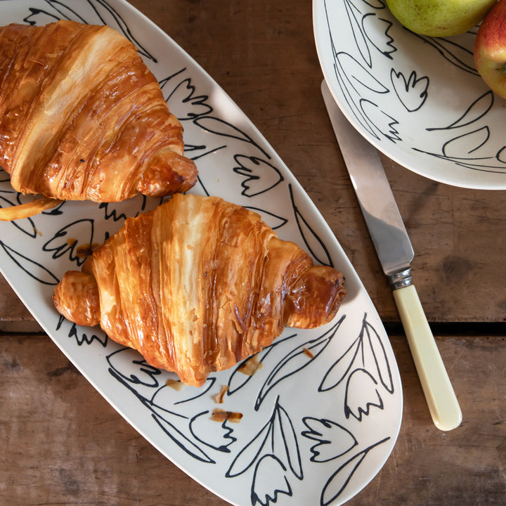 Two golden-brown croissants on a white oval serving platter with a black abstract line pattern, set on a wooden table next to a knife and another plate with fruit.