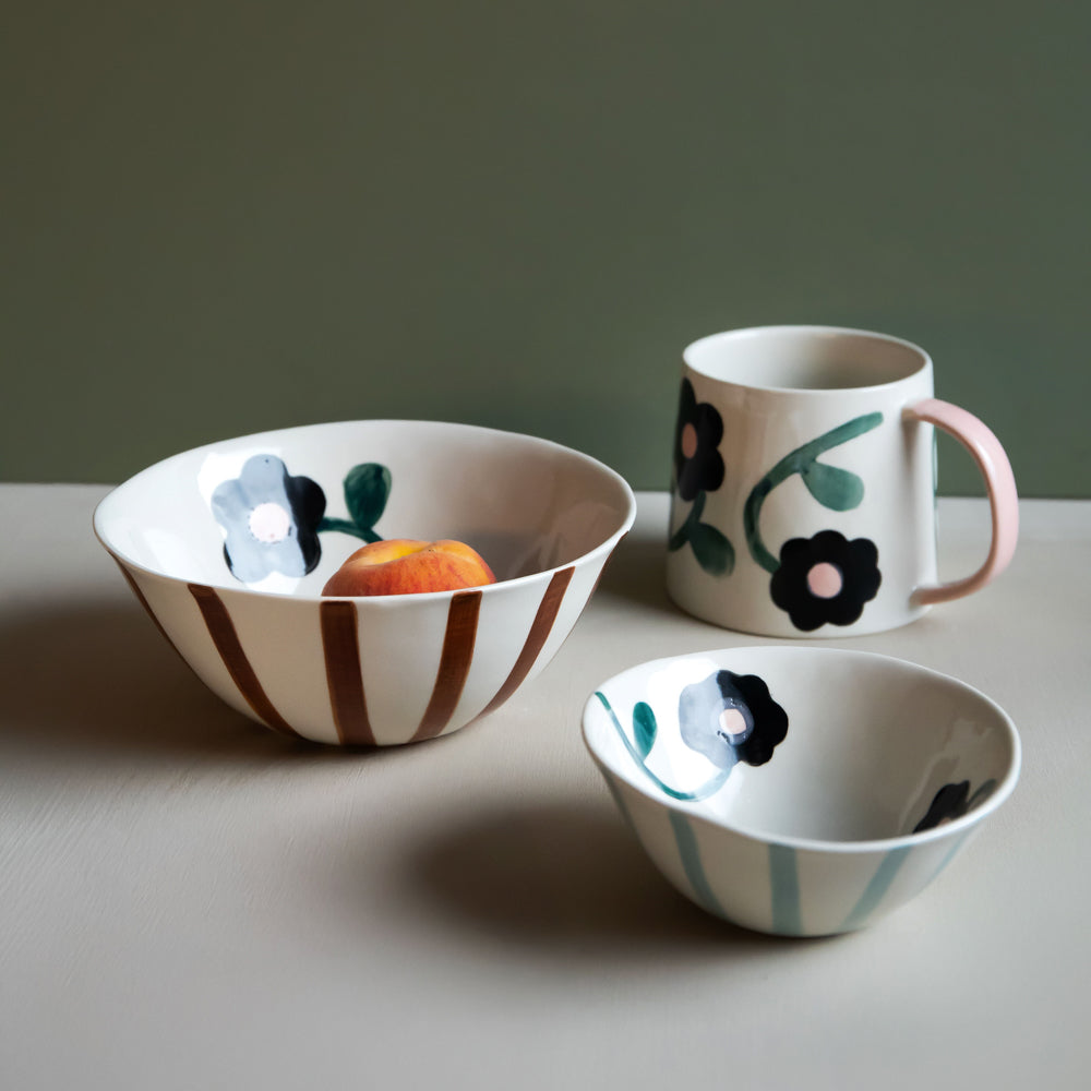 Three ceramic tableware pieces on a tabletop: a large bowl with brown vertical stripes and a peach inside, a small bowl with pale blue stripes and dark floral motifs, and a mug with a pink handle and black floral motifs with green leaves. The background is a muted green wall.