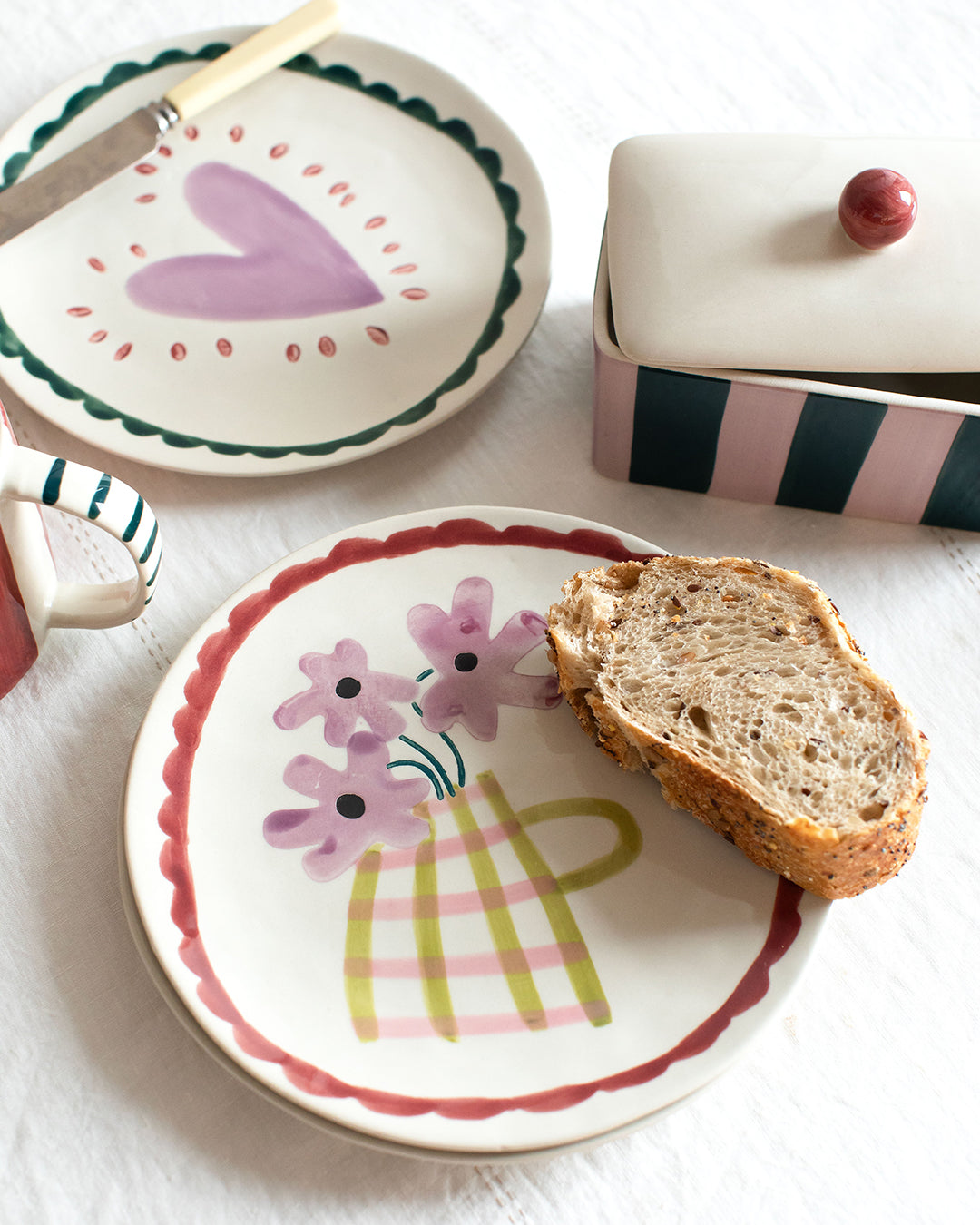 A styled tabletop scene with ceramic plates, a knife, a lidded butter box, and a slice of bread on a white fabric surface. The main visible product appears to be the decorative ceramic plate in the foreground, shown with food props rather than a person or real environment context.