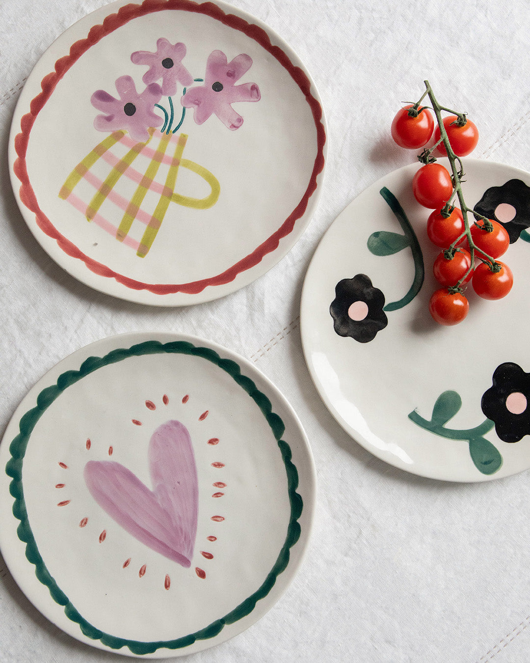 Three hand-painted ceramic plates with a rustic, folk-art style design, arranged on a white textured fabric background. One plate features pink flowers in a yellow basket, another has a pink heart with a green scalloped border, and the third displays black flowers with green leaves next to a small vine of red cherry tomatoes.
