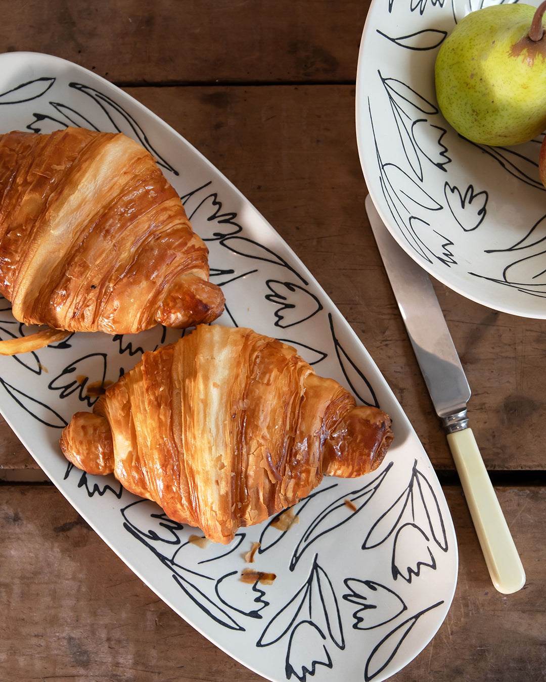 Two golden-brown croissants on a white oval serving platter with black floral line drawings, set on a rustic wooden table next to a knife and a partial plate with a pear.