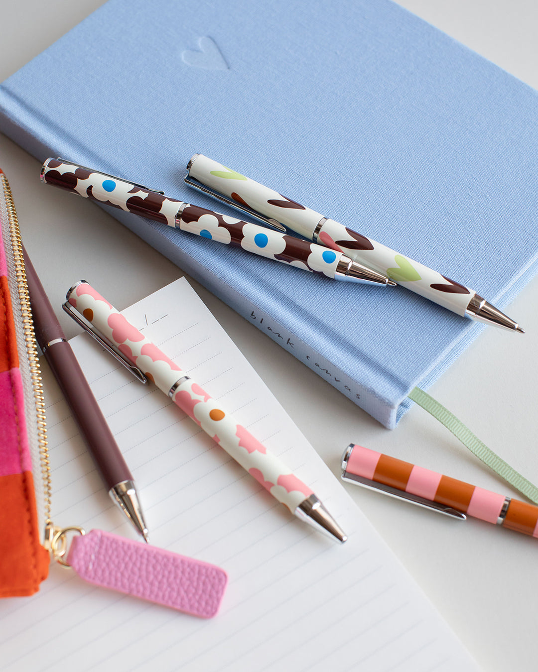 A styled flat-lay of patterned pens on a light blue notebook, with an orange zip pouch, a dark brown pen, and lined paper on a white surface.