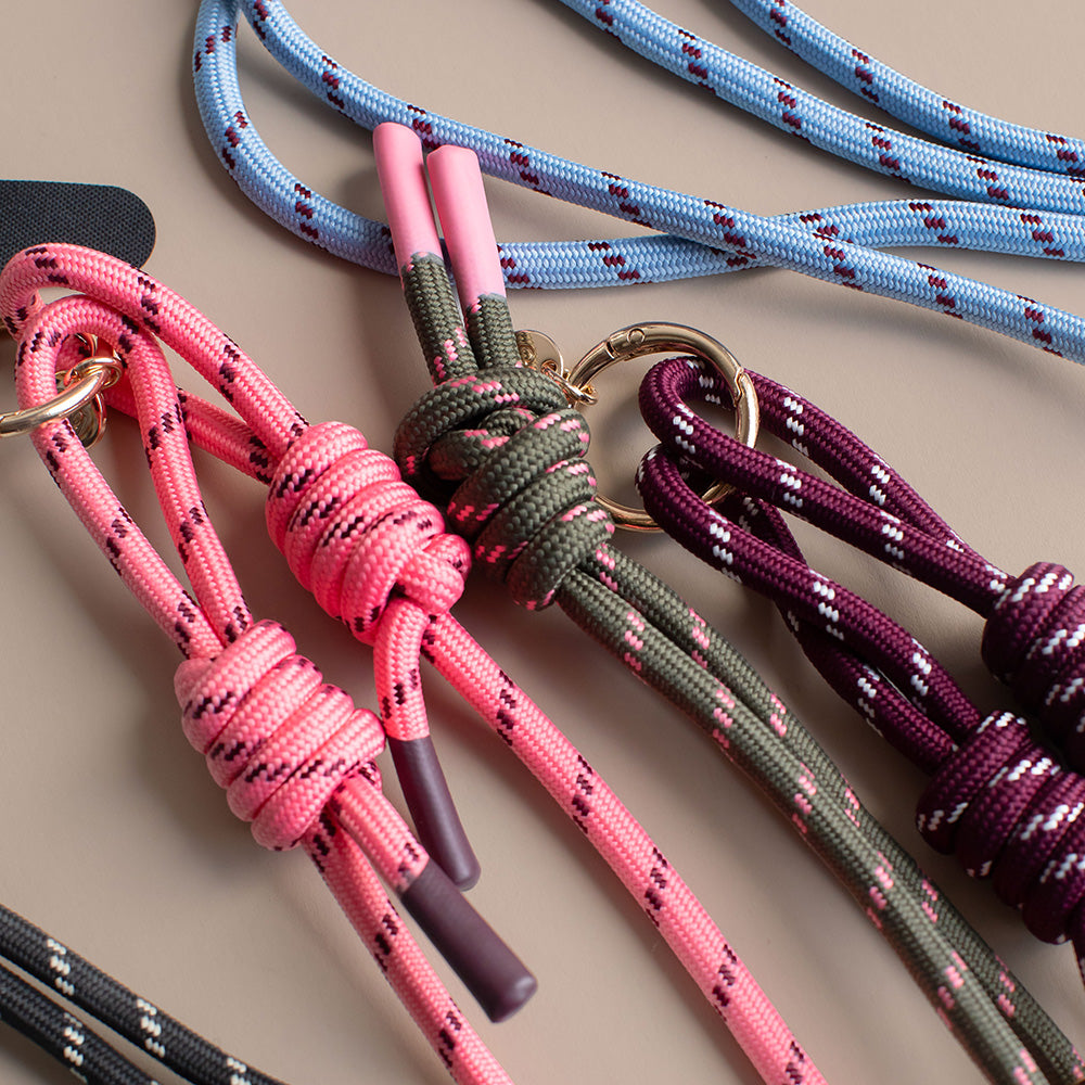 Three knotted cord phone straps in pink, olive green, and burgundy, each with metal rings and cord ends, arranged on a beige background.