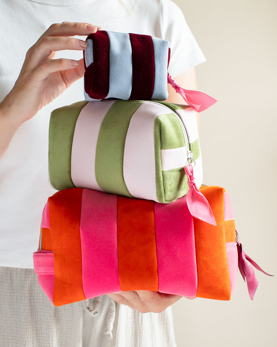 Three colorful striped fabric pouches stacked on top of each other and held by a person wearing a white shirt. The pouches have vertical color-blocked stripes in different color combinations: the top one is light blue, dark red, and white; the middle one is light green, white, and pale pink; the bottom one is orange, hot pink, and coral.