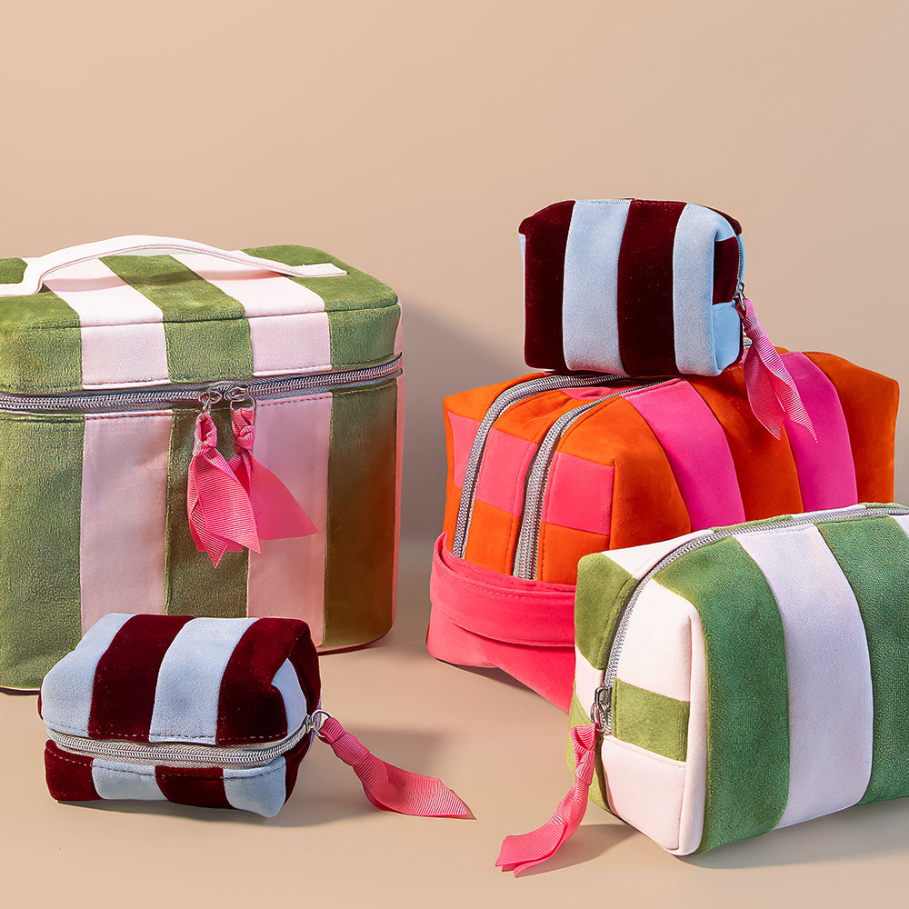 A set of colourful striped zippered wash bags & cosmetic bags and a matching vanity case made of soft velvet-like fabric, arranged on a plain beige studio background. The items feature bold vertical stripes in green, white, pink, orange, and burgundy, with matching ribbon ties.